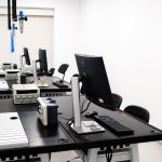 Laboratório de eletrônica com computadores, monitores e equipamentos técnicos dispostos em bancadas.